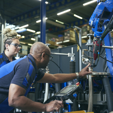 An image depicts a man and a woman collaboratively working on an advanced machine within the bustling environment of a warehouse. Both individuals are actively engaged, perhaps adjusting settings, analyzing data on a monitor, or performing maintenance, showcasing their teamwork and technical proficiency.